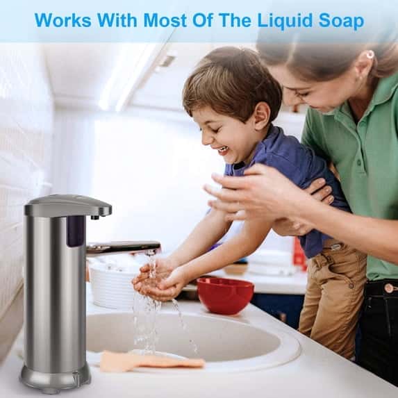 A woman helps a young boy wash his hands at a kitchen sink next to a stainless steel touchless soap dispenser.